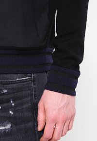 Black knit sweater with striped purple and black ribbed cuffs, textured fabric, fitted design, and visible wear on denim pants.