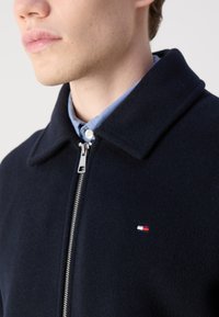 Navy wool-blend jacket with a zip front, large collar, and small embroidered logo. Buttoned light blue shirt underneath.