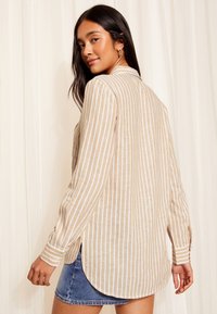 Ilgomis rankovėmis, šviesiai rudas ir baltas dryžuotas marškinėlis su išlenkta apačia ir šoninėmis kišenėmis, pagamintas iš lengvos medžiagos. Rankovių mygtukų detalė.