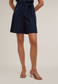 Pantaloni corti di cotone blu navy con pieghe, due tasche anteriori e vita legata, abbinati a sandali con tacco alto beige con cinturini.