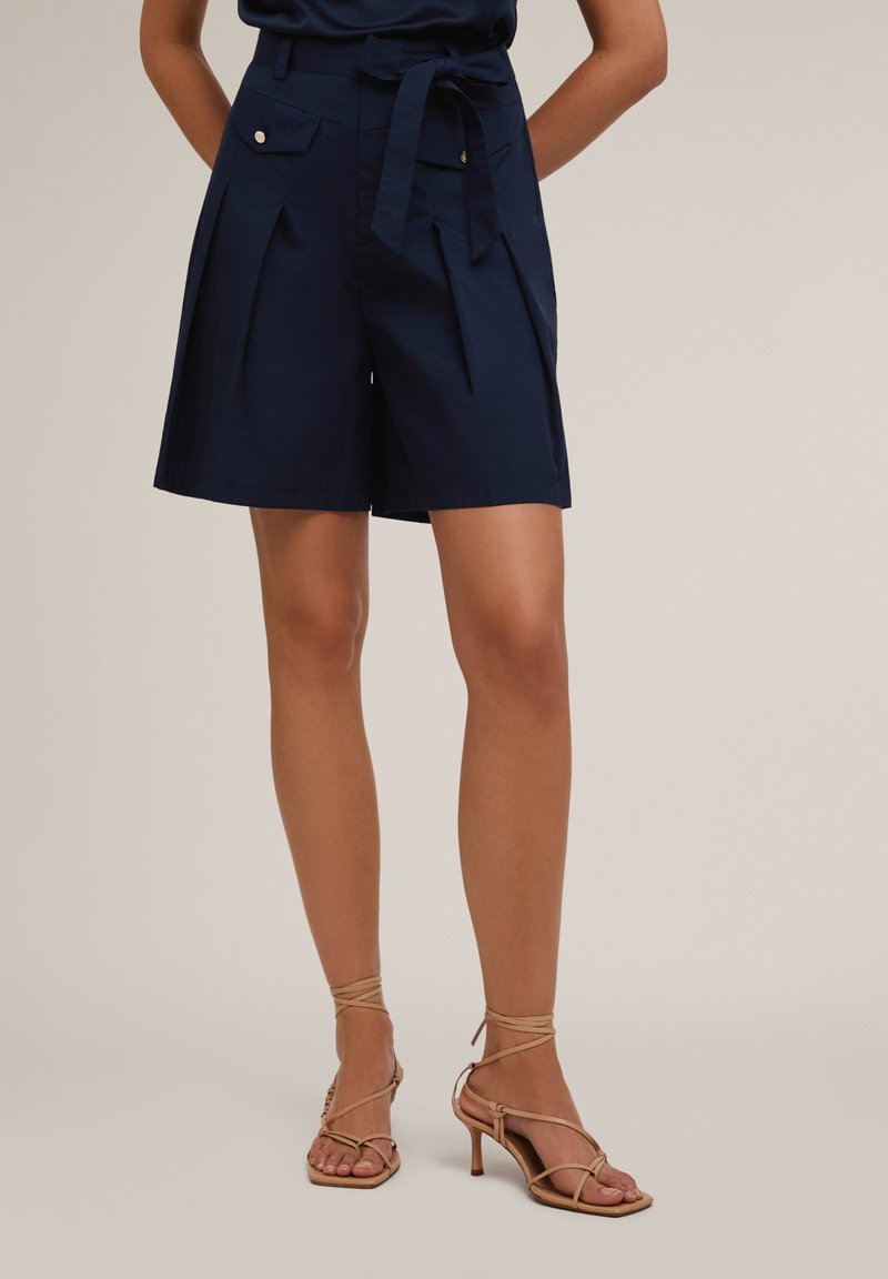 Pantaloni corti di cotone blu navy con pieghe, due tasche anteriori e vita legata, abbinati a sandali con tacco alto beige con cinturini.
