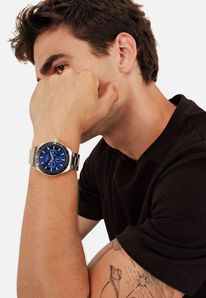 Montre en acier inoxydable argenté avec un cadran bleu et trois sous-cadrans, portée au poignet. Fond blanc, mettant en avant une chemise noire à manches courtes.