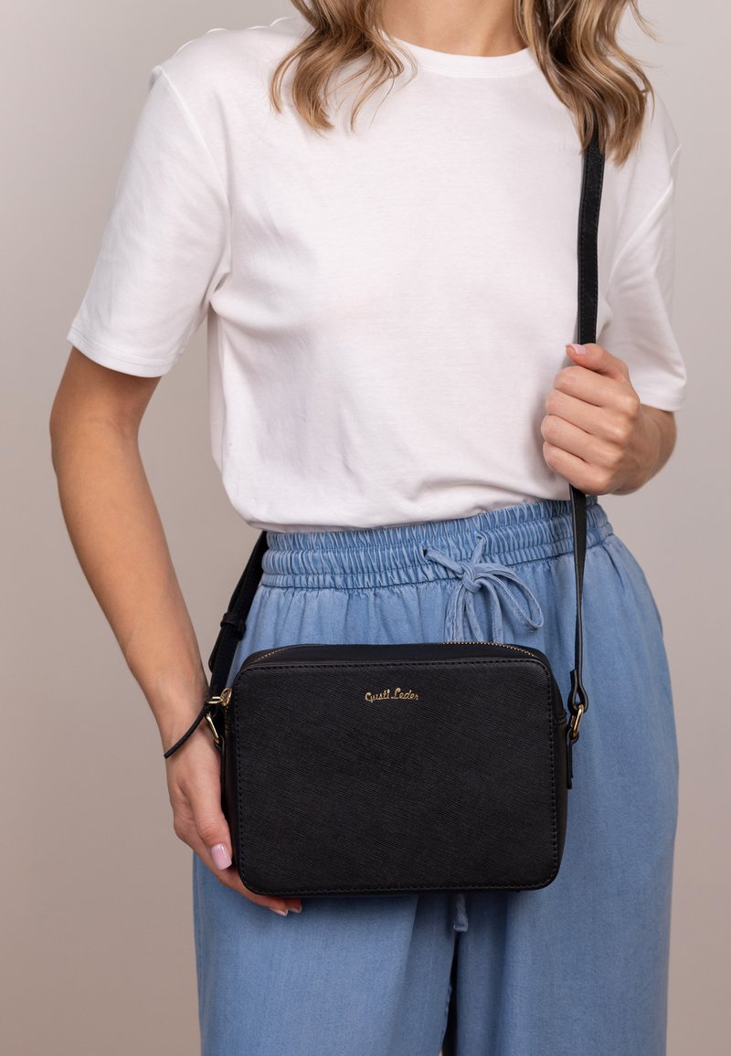 Bolso cruzado negro con una superficie texturizada, logo dorado y cierre de cremallera; combinado con un atuendo informal de camisa blanca y pantalones azul claro.