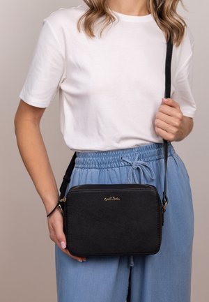 Crna torbica preko tijela s teksturiranom površinom, zlatnim logotipom i zatvaranjem na patentni zatvarač; nosi se uz ležernu odjeću bijele majice i svijetloplavih hlača.