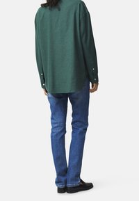 Grünes, gewebtes Hemd mit langen Ärmeln, Knopfdetails und abgerundetem Saum, kombiniert mit blauen Denim-Jeans und schwarzen Schuhen.