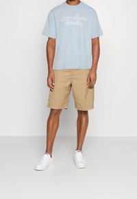 Světle modré bavlněné trikó s grafikou "Love Lights Studios", spárované s béžovými šortkami a bílými teniskami. Jednoduchý design, uvolněný střih.