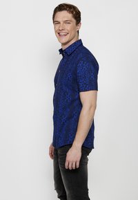 Camicia a maniche corte con bottoni in un tessuto blu a fantasia, caratterizzata da un design geometrico e bottoni a contrasto, abbinata a jeans scuri.