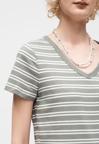 Άτομο που φοράει πράσινο και λευκό ριγέ μπλουζάκι με V-λαιμόκοψη και στρωμένα μαργαριταρένια κολιέ με μπλε λεπτομέρειες.