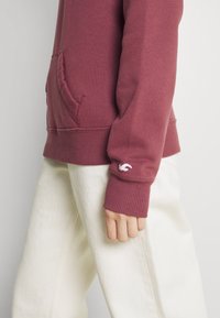 Persona vestida con un sudadera color marrón con un bolsillo frontal y pantalones blancos, con la mano relajada a un lado contra un fondo liso.