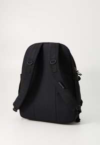 Sac à dos noir avec des bretelles rembourrées réglables, texture lisse, fermeture éclair et détail de logo sur le côté. Design simple et épuré.