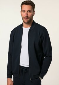 Chaqueta bomber de color navy en tejido suave con cierre de cremallera, puños acanalados y bolsillos laterales, llevada sobre una camiseta blanca.