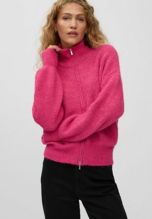 Femme aux cheveux blonds bouclés portant un pull zippé rose vif et un pantalon noir, posant avec une main près de son visage.