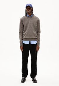 Grauer strukturierten Pullover über einem hellblau gestreiften Hemd, kombiniert mit schwarzen Cord-Hosen und schwarzen Schuhen, ergänzt durch eine blaue Mütze.