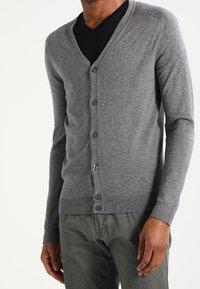 Grå kardigan av mjukt tyg, med V-ringning, femknäppning, ribbade ärmar och en slim fit-design. Bärs över en svart skjorta.