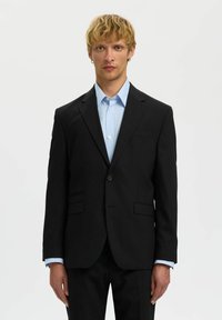 Zwarte op maat gemaakte blazer met gesneden revers, enkele knoopsluiting, twee voorzakken, gedragen over een lichtblauw gestreept shirt.