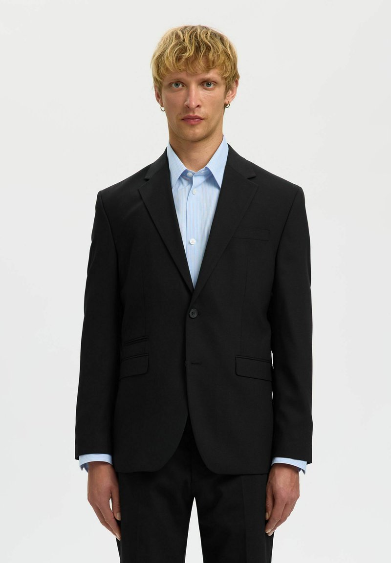 Zwarte op maat gemaakte blazer met gesneden revers, enkele knoopsluiting, twee voorzakken, gedragen over een lichtblauw gestreept shirt.