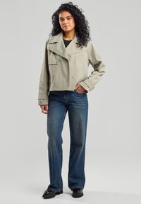 Lyhennetyttö harmaa takki, jossa on suuret läpät ja kaksiriviset napit, käytetty mustan yläosan ja klassisten sinisten farkkujen päällä. Mustat kengät täydentävät asua.