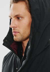 Czarna kurtka z kapturem i wysokim kołnierzem, z bordowym wewnętrznym wykończeniem, widocznymi zapięciami na napy oraz gładką, matową fakturą.