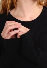 Svart ribbad tröja med långa ärmar, med strukturerad tyg och lösa ärmände, bärs på en persons överkropp och händer.