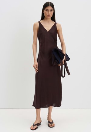 Femme aux longs cheveux foncés portant une robe midi marron foncé sans manches et des sandales à talons foncées, tenant une veste ou un sac foncé plié.