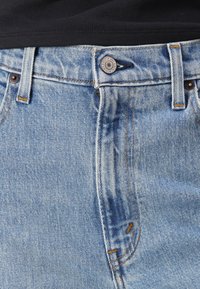 Jean en denim bleu clair avec une fermeture à bouton, des coutures en cuivre et un design classique à 5 poches. La texture est lisse avec un léger effet usé.