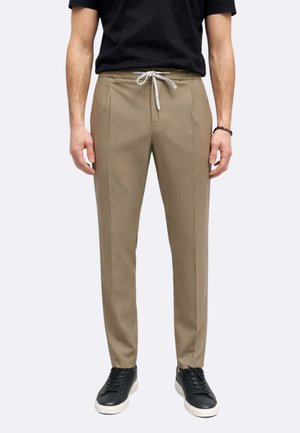 Uomo con pantaloni beige con coulisse, scarpe da ginnastica nere con suola bianca e t-shirt nera, in piedi contro uno sfondo semplice.