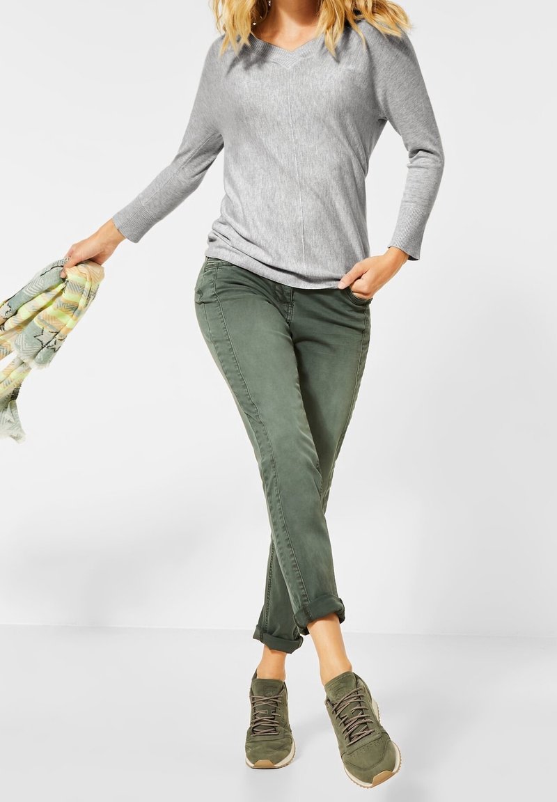 Graues Langarm-V-Ausschnitt-Shirt, olivgrüne Hosen mit umgeschlagenen Ärmeln und dunkelgrüne Schnürsneaker. Hält einen gemusterten grün-gelben Schal.