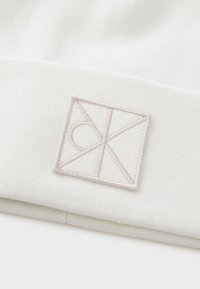 Valge kootud müts, millel on tekstuuriga helebežž embroideeritud logo geomeetrilises kujunduses. Pehme kangas ja volditud äär.