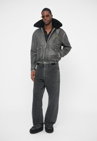 Veste en denim avec col en fourrure, couleur noire, fermeture éclair à l'avant, deux poches latérales, associée à un jean noir à jambe large et des chaussures noires à motifs.