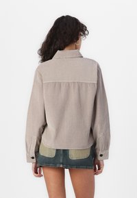 Krémová kordurová košile s límečkem, dlouhými rukávy a knoflíkovými manžetami; má zadní yoke a sklady pro pohodlný střih. Kombinováno s džínovou sukní.
