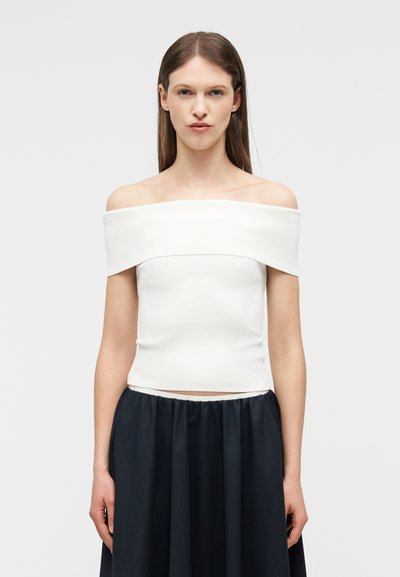 Femme aux cheveux bruns lisses portant un haut blanc décolleté et une longue jupe noire, debout devant un fond clair uni.