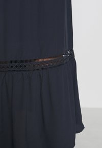 Marinblå klänning med samlad nederdel, strukturerad tyg och dekorativ spetsdetalj vid midjan med utskurna mönster.