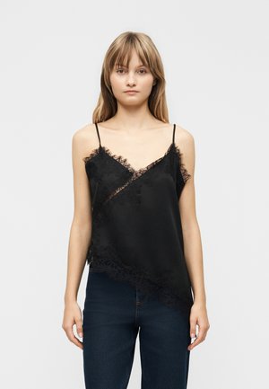 Ung kvinde iført en sort top med blonderkant og mørkeblå jeans, stående foran en ensfarvet lys baggrund.