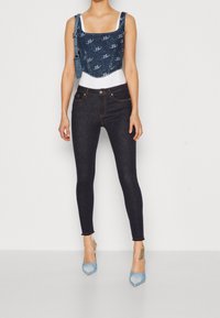 Top corto de denim con un patrón de logo, combinado con jeans ajustados oscuros. La modelo lleva zapatos de tacón de color azul claro. Diseño simple y contemporáneo.