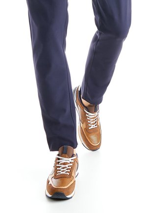 Brown sneaker with mesh and suede materials, white laces, and a navy blue pant. Features a patterned sole and brown accents.