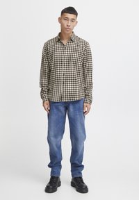 Camisa de cuadros marrón y crema con botones y mangas largas, combinada con jeans de mezclilla azul y botas negras de cordones. Fondo blanco liso.