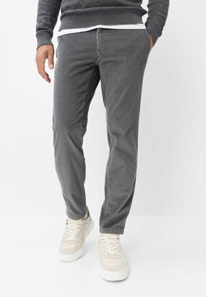 Mand iført grå kordbuks og beige sneakers, med den ene hånd i lommen og en grå sweater synlig ved taljen.