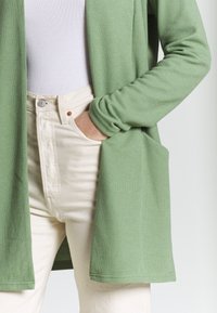 Zelený texturovaný cardigan s otevřeným předním dílem a dlouhými rukávy, kombinovaný s vysokým pasem světlých džín s předními kapsami.