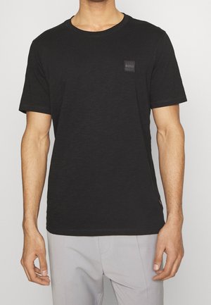 Mann trägt ein schwarzes T-Shirt mit kurzen Ärmeln und kleinem "BOSS"-Logo auf der Brust, kombiniert mit hellgrauen Hosen, steht vor neutralem Hintergrund.