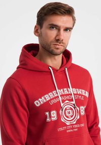 Röd hoodie med vittryckta texter och grafik. Den har en huva med dragsko och en känguruficka. Tillverkad av mjukt tyg med en avslappnad passform.