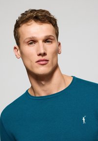 Tealfarbenes Rundhals-Sweatshirt aus weichem Stoff, mit geripptem Halsauschnitt und kleinem weißen Logo auf der Brust. Glatte Textur, enganliegendes Design.