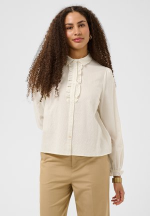 Femme aux longs cheveux bouclés portant un chemisier crème à volants et un pantalon beige, debout devant un fond clair uni.