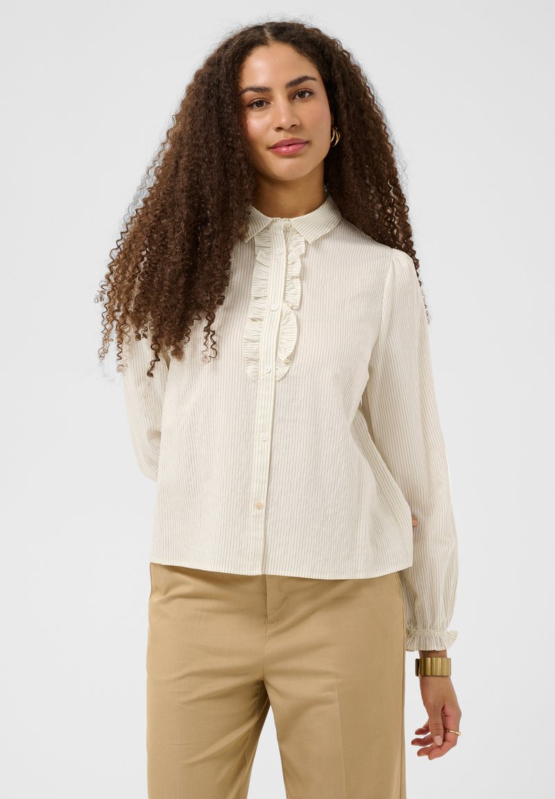 Femme aux longs cheveux bouclés portant un chemisier crème à volants et un pantalon beige, debout devant un fond clair uni.