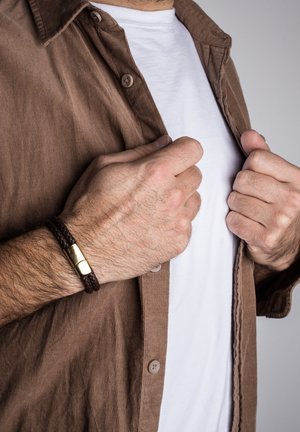 Braune Cordhemd mit einem weißen T-Shirt darunter, das ein braunes geflochtenes Lederarmband mit einem goldenen Metallschnallen am Handgelenk zeigt.