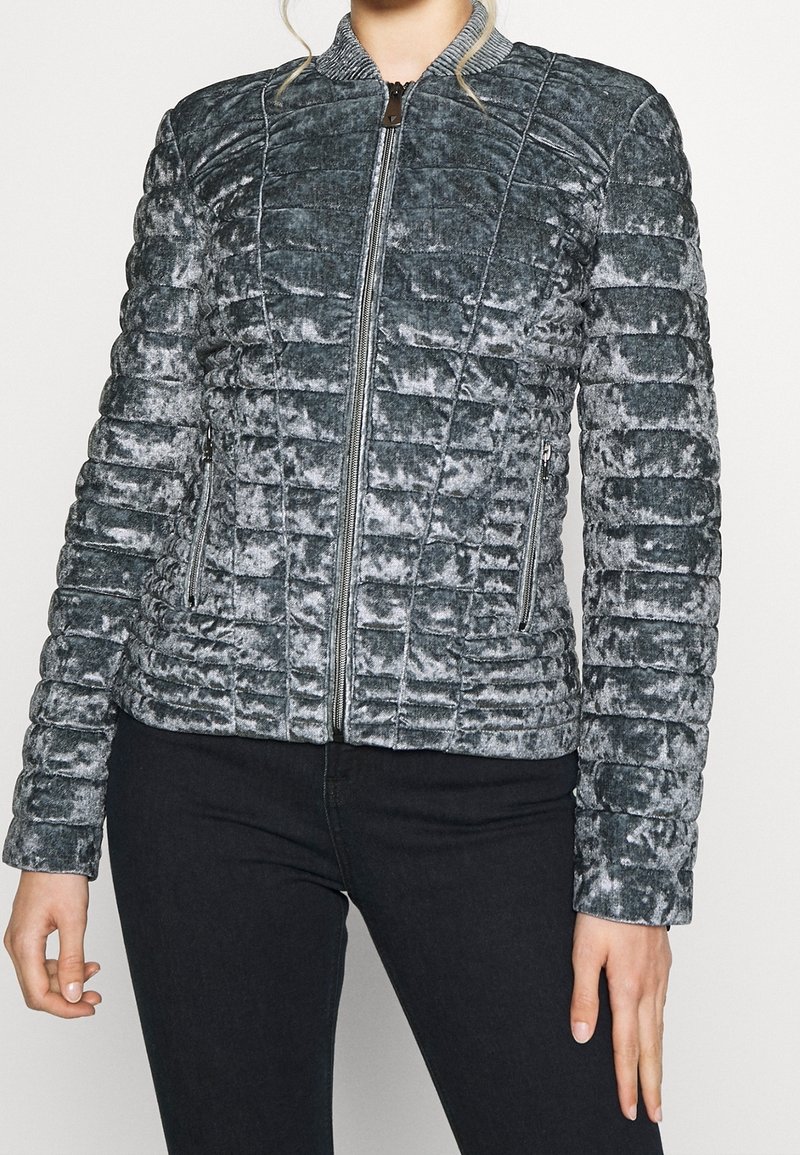 Femme portant une veste bomber en velours matelassé gris avec une fermeture éclair à l'avant et des poches zippées, associée à un pantalon noir.