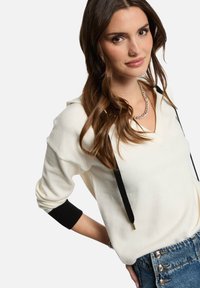 Felpa bianca con dettagli neri e coulisse. Presenta un collo a V e maniche arrotolate. Indossata con jeans a vita alta e bottoni decorativi.