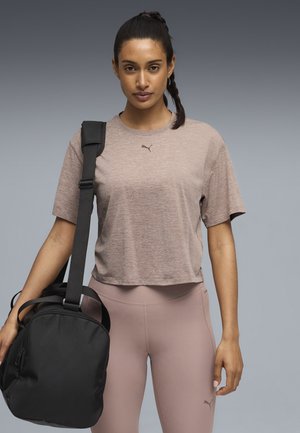 Lichtbruine cropped t-shirt met korte mouwen en een subtiel logo, gecombineerd met roze leggings. Een zwarte sporttas hangt over een schouder.