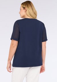 Mujer con cabello rubio lleva una blusa de manga corta azul marino con mangas transparentes y pantalones blancos, vista desde atrás contra un fondo liso.