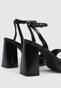 Sandalias de tacón negro con tacón cuadrado y correas elegantes, con un acabado brillante y hebilla ajustable en el tobillo para un ajuste seguro.