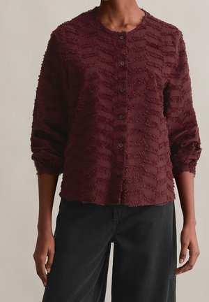 Femme portant un chemisier bordeaux texturé boutonné avec manches longues et un pantalon noir, debout devant un fond neutre.